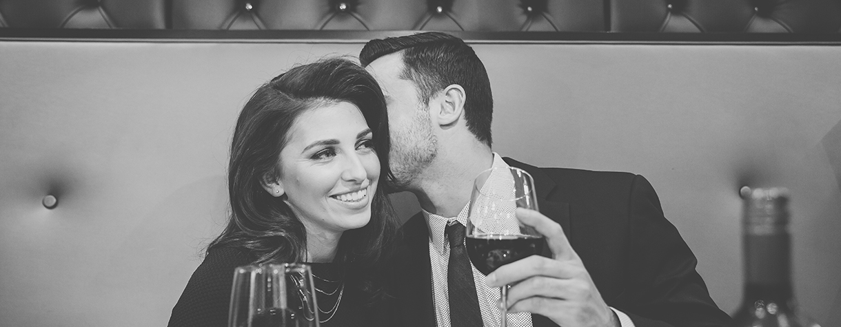 Man holding a glass of wine and whispering into a woman's left ear